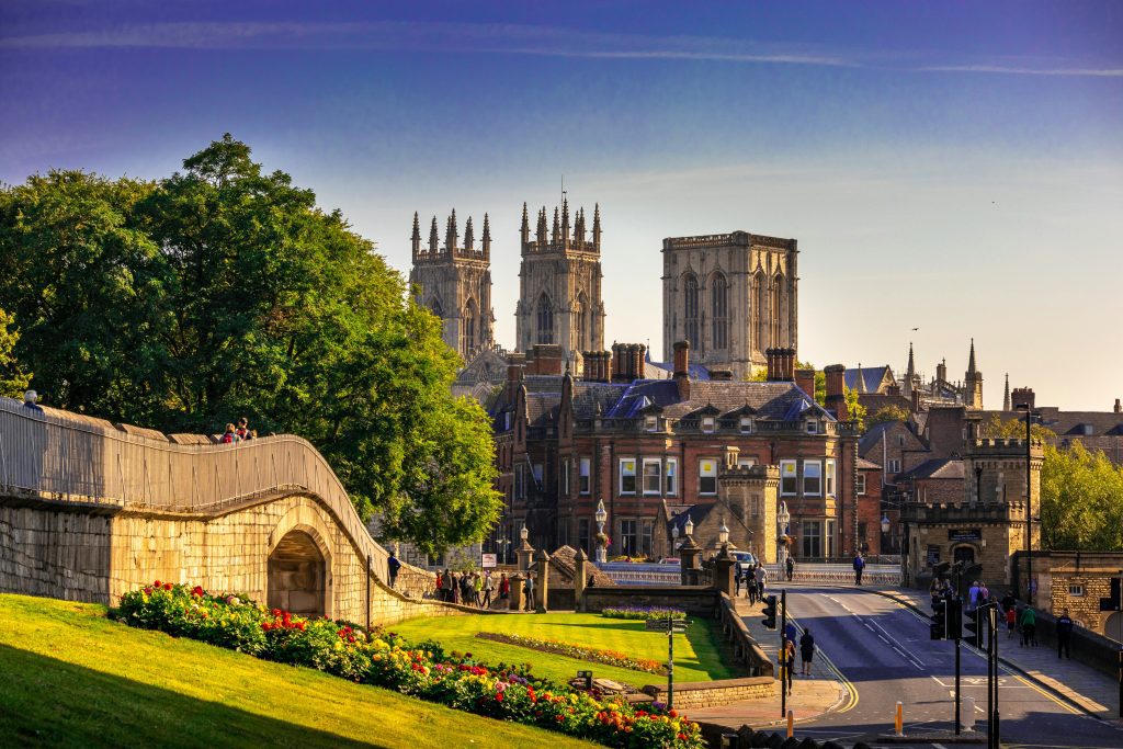 A sunlit area of a town with grass and flowers, a street and some buildings, with a cathedral looming in the background